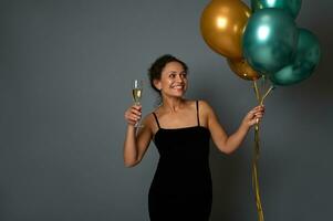 Pretty woman with shiny golden green metallic air balloons posing against gray wall background with copy space holding a glass of champagne in hands. Attractive lady celebrates festive event photo