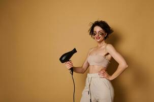 Waist-length portrait of stylish European woman with smoothing collagen patches under eyes, in beige underwear blowing hair dryer, dries short wet hair on beige background with copy space photo