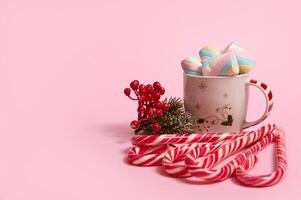 Beautiful minimalistic Christmas composition with hot chocolate drink decorated with marshmallows and striped lollipops, candy canes isolated over pink colored backgroud with copy space for ad photo