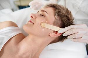 Closeup of aesthetician hand using wooden spatula for applying transparent anaesthetic gel on woman's face for making the laser removal hair procedure non painful. photo