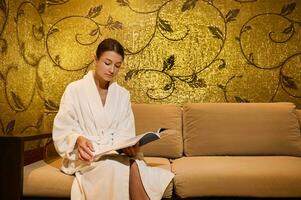 Confident well-groomed middle aged woman in white terry bathrobe reading magazine sitting in a couch in a lounge area of a luxury wellness spa centre. Professional body, skin care, relaxation concept photo