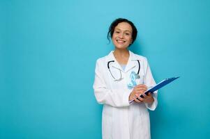 Middle aged female mixed race doctor in white medical gown, wears a blue awareness ribbon, poses with a clipboard in her hands, cutely smiled looking at camera,copy space. World Diabetes Awareness Day photo