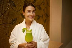 Middle aged European woman in white bathrobe, holding a glass with healthy freshly squeezed vitamin juice in her hands, smiles toothy smile looking at camera, standing in wellness centre lounge area photo