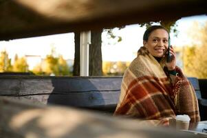 View through wooden logs of bench at beautiful woman looking to the side while talking on cell phone, sitting on wooden bench wrapped in cozy checkered woolen blanket keeping warm on cool autumn day photo