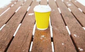 Yellow takeaway cup of hot drink, coffee or tea on wooden bench in a snowy winter park photo