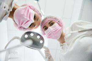 Bottom view of female doctor dentist and assistant adjusting medical light lamp, holding dental instruments and leaning toward a patient for medical examination in white modern clinic. Dental practice photo