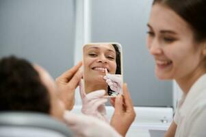 de cerca de medio adulto mujer participación cerca su dientes un color gráfico para blanqueamiento dientes y comparando el blancura de dientes con dental muestra, sonriente con dientes sonrisa mirando a su espejo reflexión foto