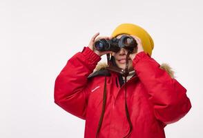 Handsome school boy in yellow woolen hat and bright red down jacket, smiles toothy smile looking through binoculars, isolated on white background with copy space. Winter travel and adventure concept photo