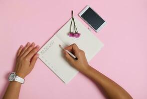 Top view of female hands holding pencil, writing in notebook, checking to-do list. Mobile phone and pink flower lying on pink background with copy space. Business, planning and time management concept photo