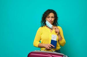 Sad young woman in bright yellow sweater takes off her protective medical mask and sadly looks at camera, posing against blue background, holding suitcase, air ticket and boarding pass with passport photo