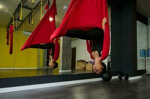 Young sporty African woman practicing aerial flying yoga stretching exercises, being in bat pose while hanging on red hammock photo