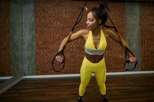 Beautiful slim body African woman with flat belly doing arm exercises and push-ups while working out with suspension straps at gym. Healthy and active lifestyle, body care, fitness and sport concept photo