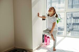 Beautiful little girl standing near a wall and learning performing tree pose in yoga. photo