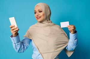 Young woman in hijab holding a white blank plastic card in her hand and texting on mobile phone in her other hand photo