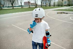 Upset boy skateboarder in protective gear frustratedly wipes tears from his eyes and holds a skateboard in one hand while standing on a sports playground photo