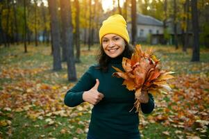 linda mujer en amarillo de lana de punto sombrero y verde pull-over sostiene un recogido hermosa seco ramo de flores de caído arce hojas y muestra un pulgar arriba, sonriente con dientes sonrisa mientras relajante en un otoño parque foto