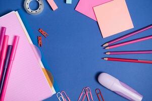 Assortment of school office accessories in pink shades, isolated on blue background, copy space. Copybook, felt tip pens, markers, paper clips,pencils and stationery supplies scattered in a circle photo