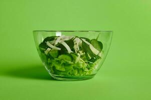 Close up of green salad with herbs, lettuce, cabbage and cucumber in transparent glass bowl on green background photo