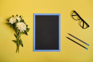 Flat lay composition from arranged aster flowers next to a chalkboard with copy space , eyeglasses and two pencils, isolated on yellow background photo