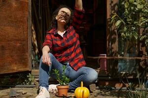 sonriente mujer cubre sí misma con su mano desde el del sol rayos que cae en su cara y disfruta jardinería mientras sentado a el peldaño en un de madera Kiosko foto
