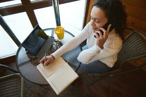 Top view of a negotiating woman by phone and writing on a diary while sitting near window at a wooden cafe photo