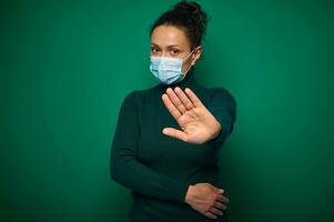 Isolated portrait on green background of a serious millennial woman in protective medical mask, gesturing, showing stop sign with her hand looking at camera. Copy space photo
