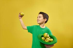 Funny little boy stands on yellow background and holds an Irish green cap full of apples in one hand and looks at a green apple in the other. Saint Patrick's day photo