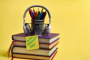 Composition from a stack of colorful books pencils glasses wireless headphones note paper with lettering Teachers Day on a yellow background with copy space. Back to school concepts. photo