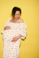 Multi-ethnic body positive charming delightful pregnant woman expecting a baby, holding belly, isolated on yellow photo