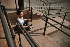 joven latín americano mujer en auriculares utilizando móvil teléfono mientras sentado en patineta en antecedentes de pasos foto