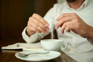 Close-up of an unrecognizable man in a white shirt sprinkling sugar from sticks into a white ceramic cup with a freshly brewed coffee drink, sitting at a wooden table and enjoying a coffee break photo