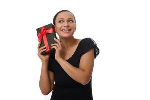 Curious beautiful mixed race woman dressed in black holds a gift box near her ear and listens to what it contains, smiles toothy smile, isolated over white background with copy space photo