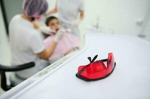 Focus on red safety glasses, protective goggles for patient used during teeth whitening in dental clinic, against blurred background of pediatric dentist and assistant treating child's oral cavity photo