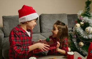Cute European kids, a boy in Santa's hat and a charming baby girl sit at a table with freshly baked Stollen bread and a hot drink with marshmallow and give each other gifts for Christmas. photo