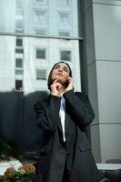 Beautiful self-confident thoughtful young woman, freelance, entrepreneur, copywriter talking on cell phone on modern office corporate high-rise buildings background. Business, communications concept photo