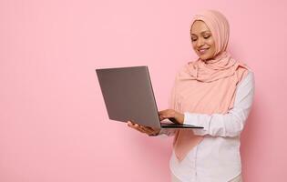 Portrait of a beautiful Muslim Arab woman in pink hijab working on laptop, isolated on colored background with copy space. Successful programmer, IT female worker, start-up, business lady photo