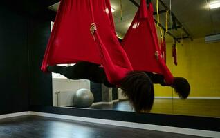 Young sporty African American slim body woman practicing aerial flying yoga stretching exercises, being in bat pose while hanging on red hammock photo