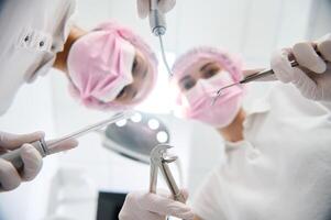 Focus on stainless steel dental instruments- extractor forceps and probe, saliva ejector and irrigator in the hands of a dentist and assistant leaning towards a patient during routine dental check-up photo