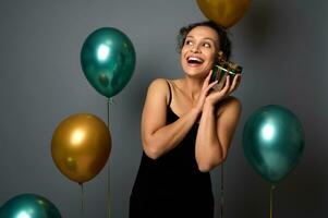 Attractive woman in evening dress holds a Christmas gift near her face, gently hugs it, looks up on copy space on gray background with gold green air balloons. New Year, Birthday party concepts for ad photo