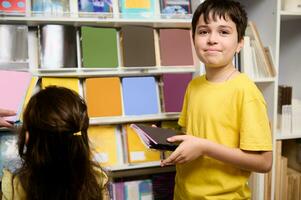 Happy adorable school kids, cute teenage boy and preschool girl choosing copybooks on shelves in school stationery store photo