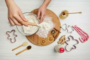 Female hands holding a spoon with flour above a bowl on a wooden table with dried orange slices and cutting molds photo