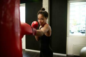 African athletic woman with perfect physique , boxer in red boxing gloves, trains hard, strikes a straight punch, hits a huge punching bag in a boxing gym. Martial art concept. photo