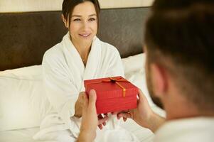 Birthday, marriage anniversary concept. Loving married couple giving each other gifts for Valentine's Day. Beautiful woman getting a red gift box sitting dressed in bathrobe on the bed in bedchamber photo