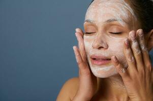 hermosa mujer masajear su cara mientras quitando maquillaje utilizando un espuma limpieza cosmético producto, y refrescante su piel con un exfoliante belleza producto, aislado terminado gris antecedentes. de cerca. foto