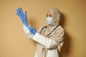 Muslim female doctor in hijab wearing medical protective gloves posing in front of camera on beige background with copy space photo