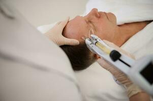 Close-up of a handsome senior man on massage table during mesotherapy gun procedure in modern wellness spa clinic. Anti-aging, rejuvenating and alternative beauty treatments to prevent first wrinkles photo