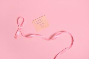 Flat lay of a long pink ribbon with endless one end, and a paper note with lettering 1 October Breast Cancer Awareness Day , isolated on pink background with space for text photo