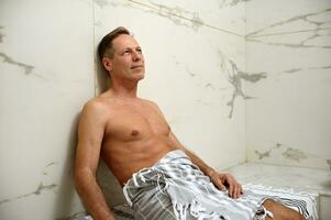 Handsome relaxed middle aged European man wrapped in a towel sitting on a marble surface in Turkish bath at wellness spa center. Body and skin care, purity, relaxation concept photo