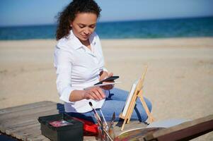 Beautiful woman sitting on a chaise lounge on the beach with smartphone in her hand. Painting tools on the foreground. photo
