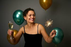 alegre dama teniendo divertido a aniversario fiesta, Navidad, nuevo años evento. hermosa mujer participación vaso de espumoso vino y bengalas, en gris antecedentes con dorado y verde aire globos foto